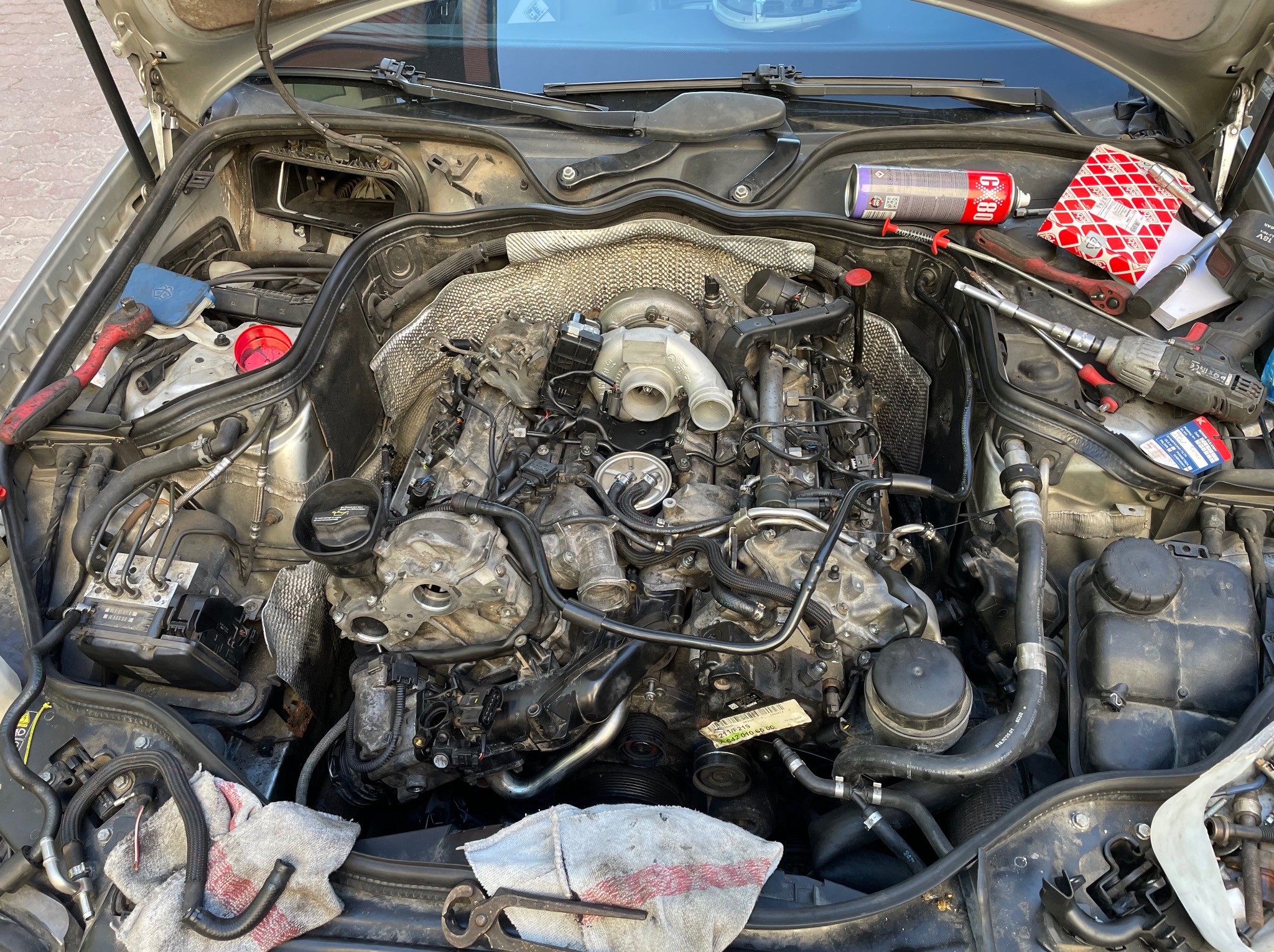 Otwarta maska samochodu z widocznym silnikiem podczas naprawy, narzędzia i akcesoria warsztatowe leżące na obudowie silnika, widoczne ślady użytkowania.