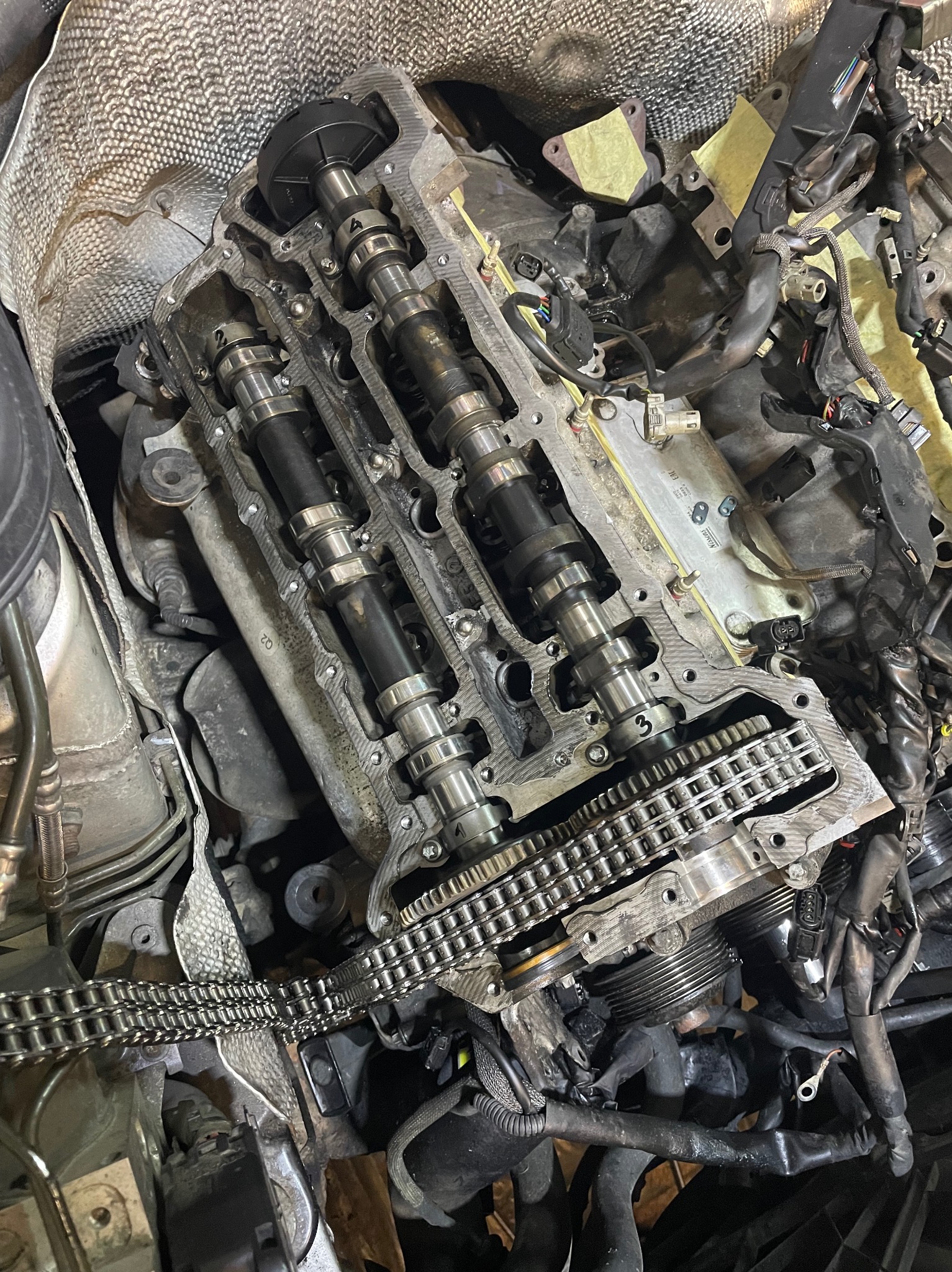 Widok z góry na otwarty silnik samochodowy z widocznymi wałkami rozrządu, łańcuchem rozrządu i przewodami w warsztacie.