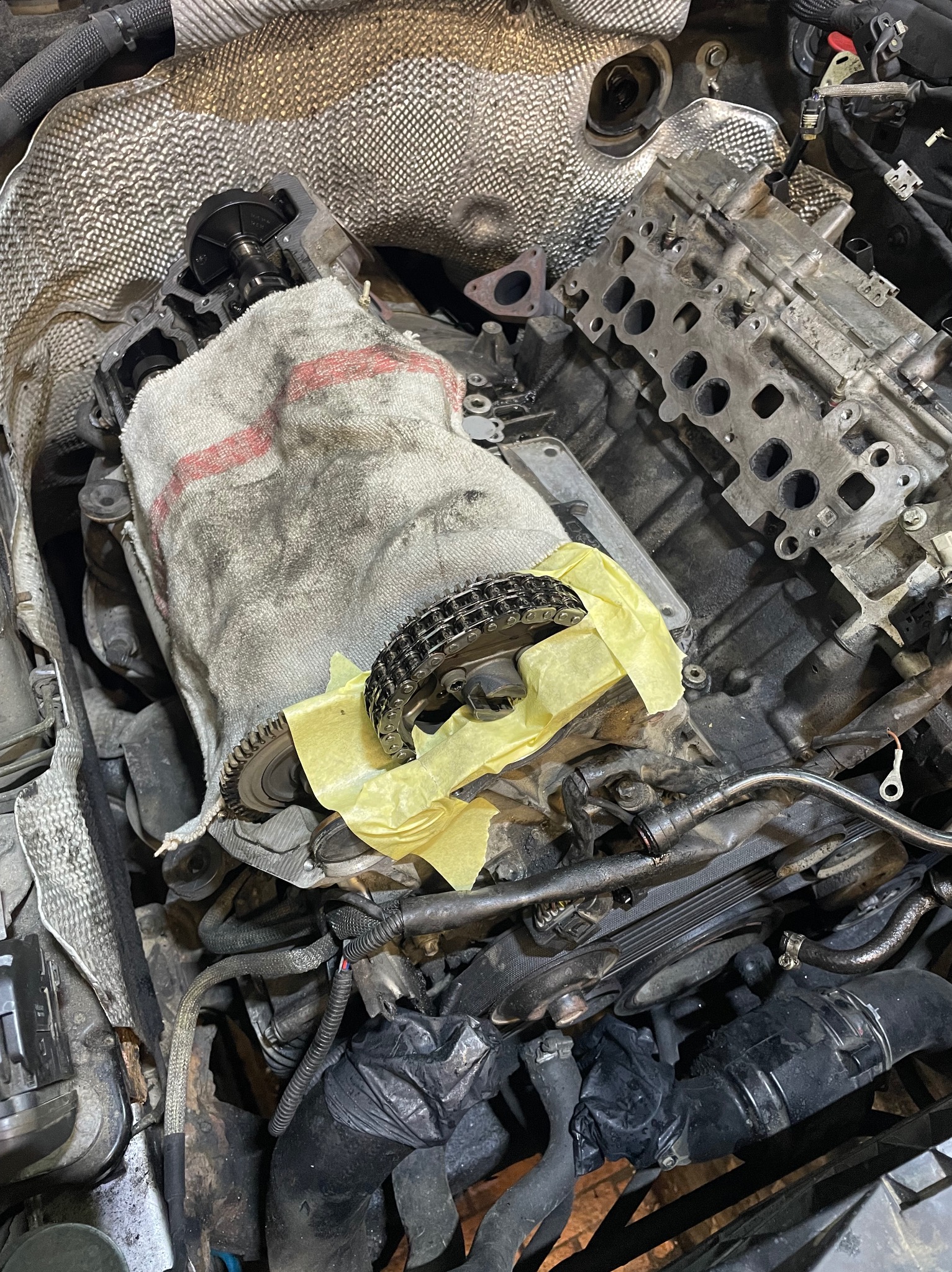 Silnik samochodowy w trakcie naprawy, widoczne elementy układu rozrządu, łańcuch i zębatka na silniku przykrytym szmatką, zabezpieczone żółtą taśmą, zdemontowana głowica silnika obok.