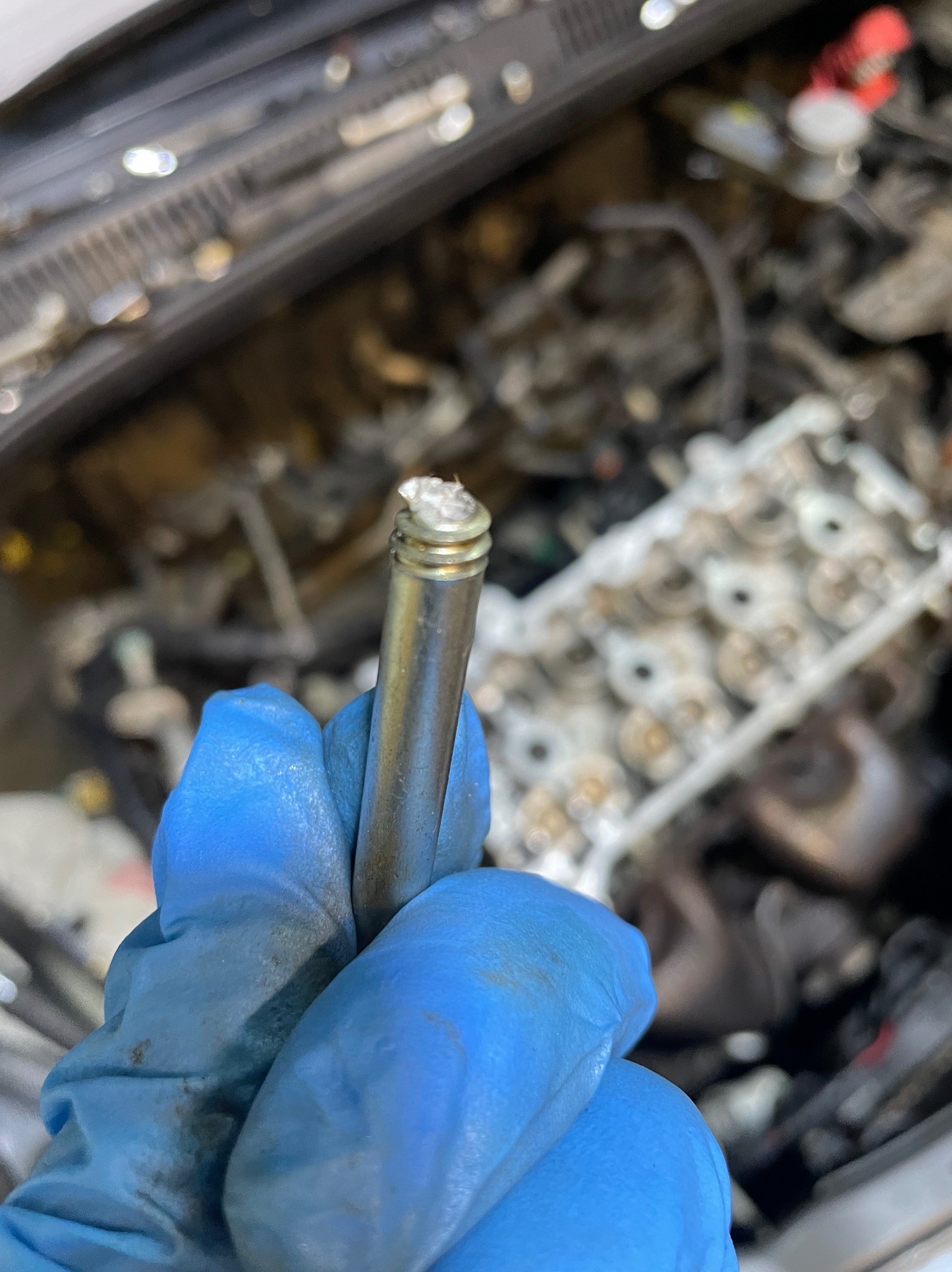 Zużyty zawór samochodowy w dłoni w niebieskiej rękawicy, z widocznymi nagarami i zabrudzeniami, na tle silnika samochodowego.