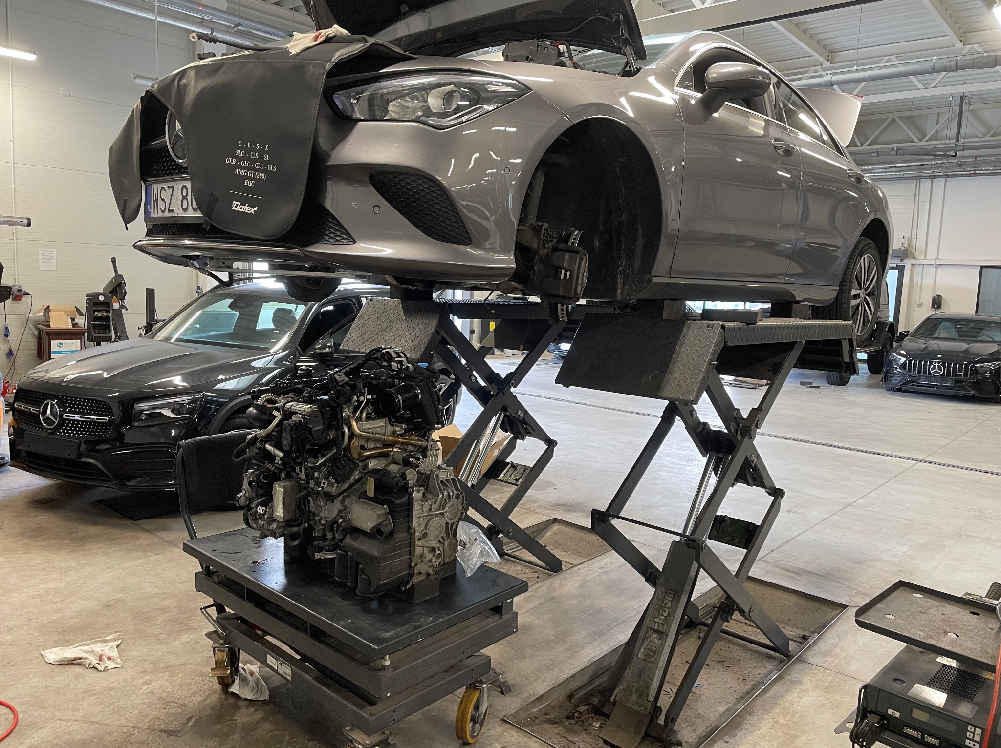 Szary samochód marki Mercedes na podnośniku warsztatowym, z zdjętym kołem i osłoną na masce, obok silnik na wózku w warsztacie samochodowym.