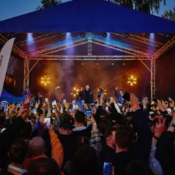 Koncert plenerowy z oświetloną sceną, tłum fanów nagrywających występ telefonami, widoczny baner reklamowy z lewej strony sceny.