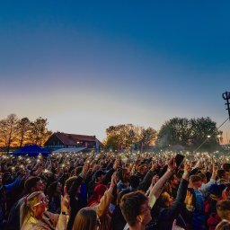 DM Event - Tłum ludzi na koncercie plenerowym wieczorem, oświetlony światłami telefonów, z widoczną sceną i budynkiem w tle.
