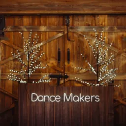 Drewniana scena z neonowym napisem 'Dance Makers', oświetlona dwoma reflektorami i ozdobiona drzewkami z lampkami, na tle rustykalnych drewnianych drzwi.