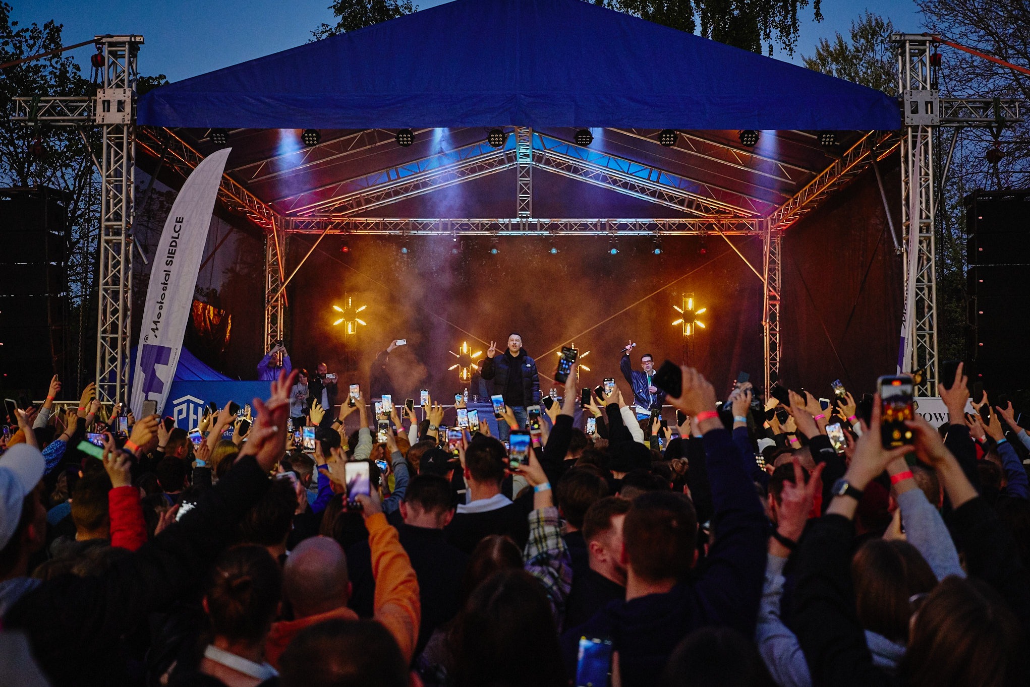 Koncert plenerowy z oświetloną sceną, tłum fanów nagrywających występ telefonami, widoczny baner reklamowy z lewej strony sceny.