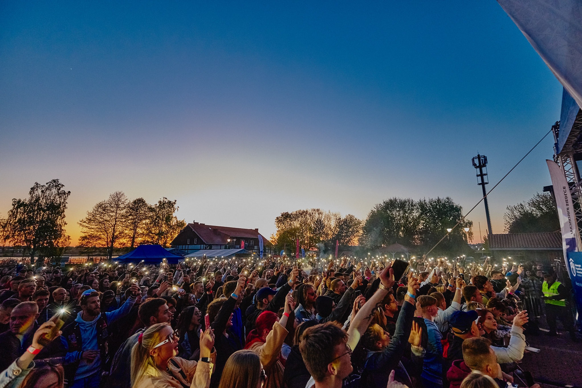 Tłum ludzi na koncercie plenerowym wieczorem, oświetlony światłami telefonów, z widoczną sceną i budynkiem w tle.