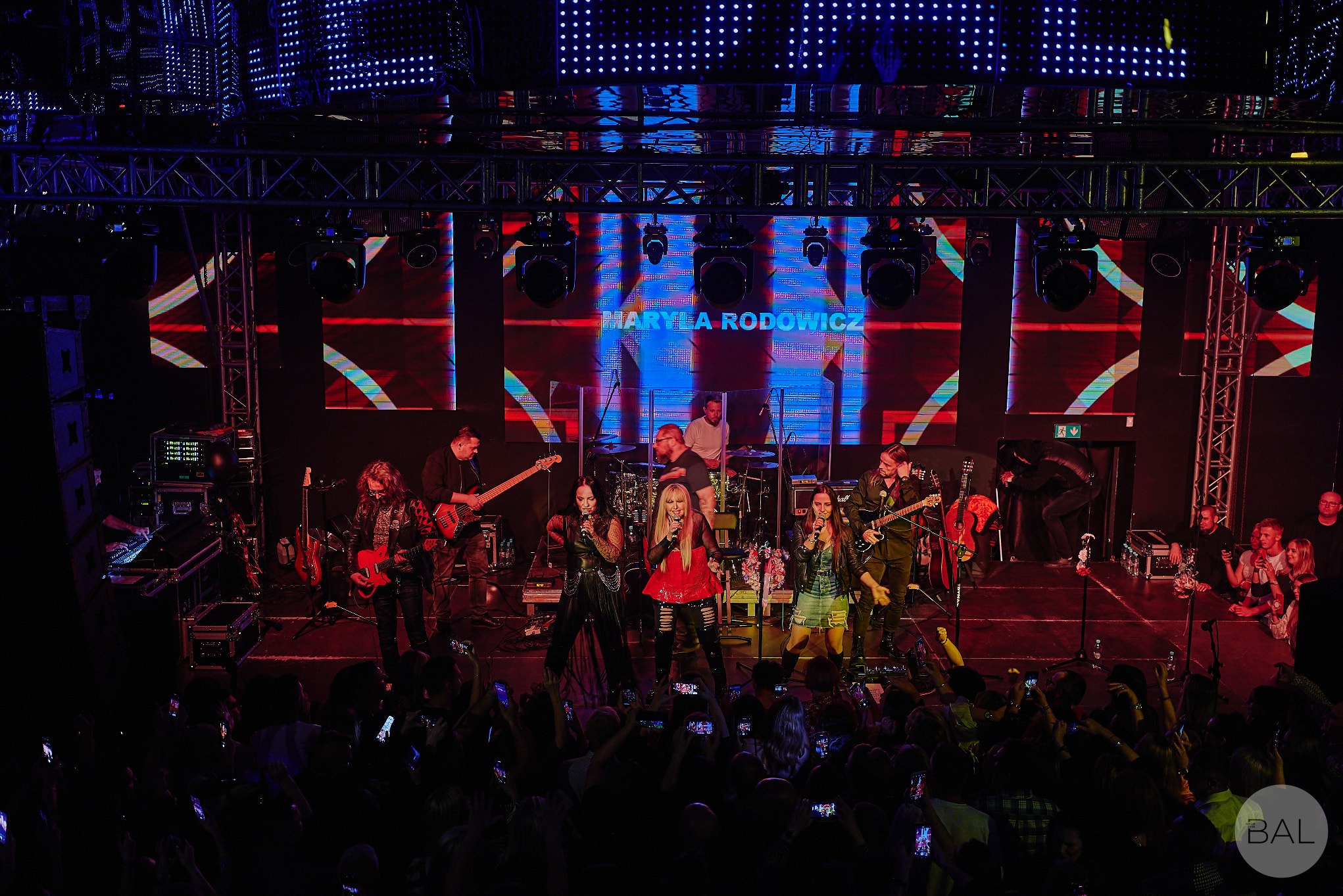 Koncert Maryli Rodowicz na scenie z zespołem, widok z dołu na publiczność z telefonami, dynamiczne oświetlenie sceniczne i telebimy w tle.