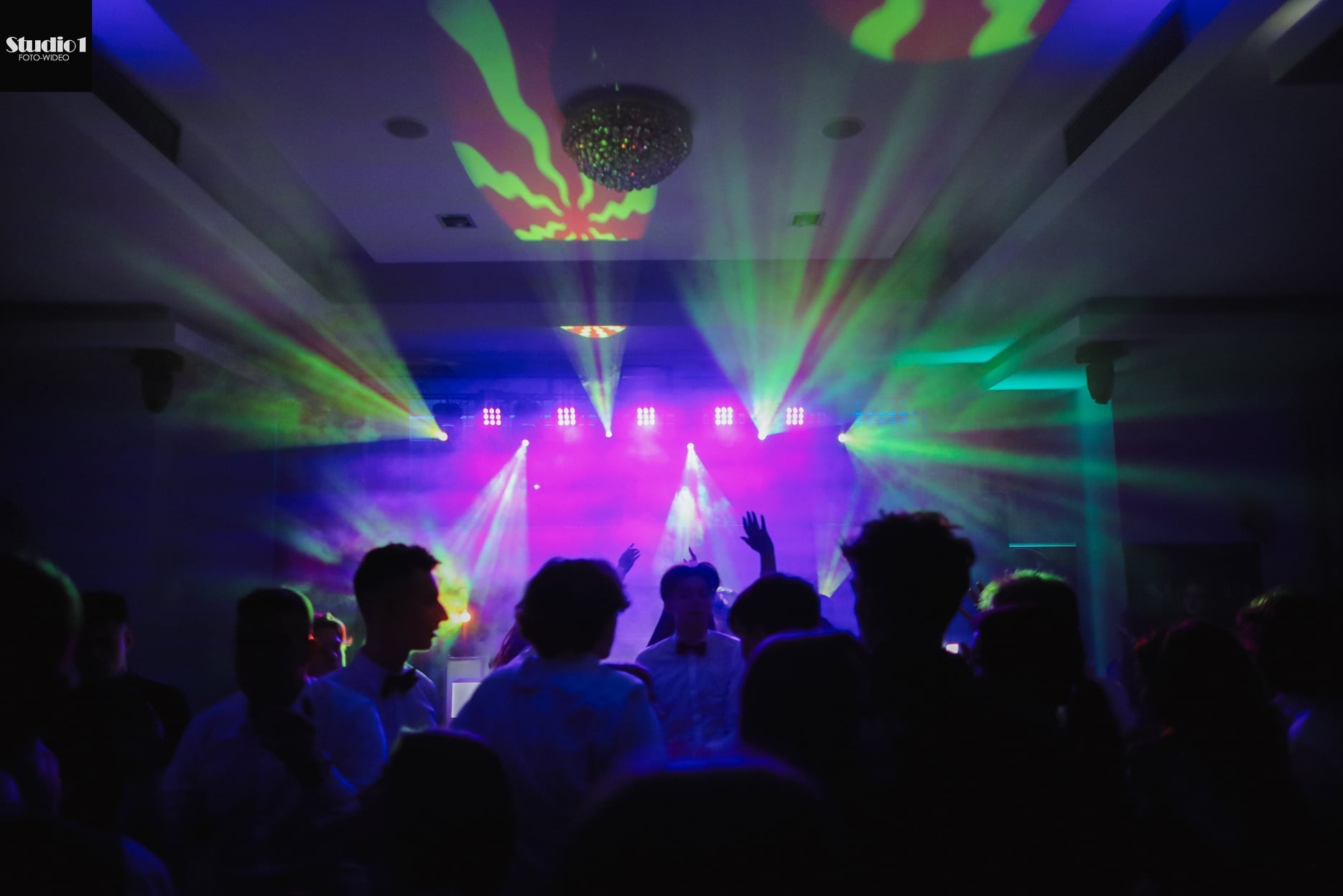 Parkiet taneczny wypełniony bawiącymi się osobami, oświetlony dynamicznymi, kolorowymi światłami scenicznymi i dyskotekową kulą.