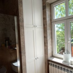 Piotr Krzyżanowski - Wysoka, biała szafa wnękowa z czterema drzwiczkami i srebrnymi uchwytami, umieszczona w łazience z brązowymi płytkami, obok okna z widokiem na zieleń.