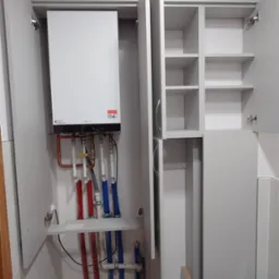 Szara szafa zabudowana z otwartymi drzwiczkami, ukrywająca piec grzewczy i rury instalacyjne. W szafce po prawej stronie widoczne półki.