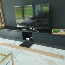 Piotr Krzyżanowski - Nowoczesna szafka RTV w kolorze grafitowym z blatem, na której stoi telewizor i ozdobna szkatułka z motywem lilii wodnych, w tle ściana z drewnianymi lamelami, na marmurowej podłodze leżą panele...