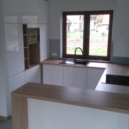 Piotr Krzyżanowski - Nowoczesna kuchnia z białymi, lakierowanymi frontami, drewnianym blatem i barkiem, czarnym zlewozmywakiem pod oknem z widokiem na ogród.