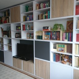 Piotr Krzyżanowski - Duża, biała szafa na wymiar z półkami, wypełniona książkami, dekoracjami i telewizorem w salonie z ciemną ścianą w tle.