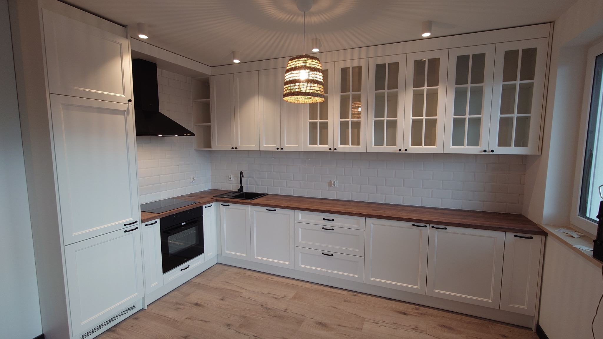 Jasna kuchnia z białymi frontami szafek, drewnianym blatem i czarnymi uchwytami, oświetlona lampą z abażurem z naturalnego materiału, z widoczną płytą indukcyjną i piekarnikiem.