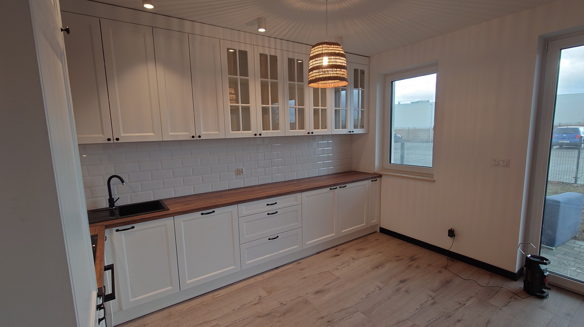 Jasna kuchnia z białymi szafkami, drewnianym blatem i lampą z wikliny. Widoczna czarna bateria i zlew, oraz okno z widokiem na zewnątrz.