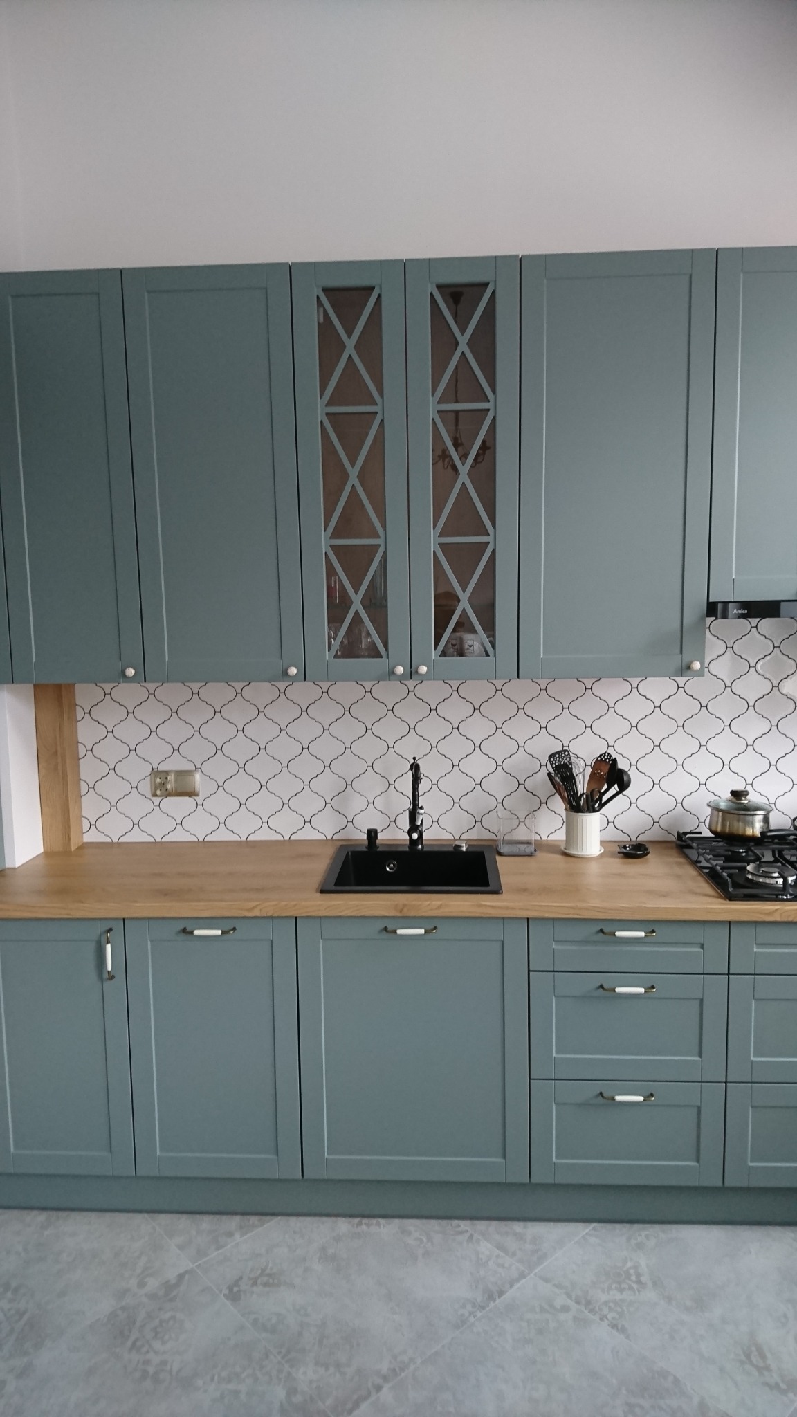 Niebiesko-szara zabudowa kuchenna z drewnianym blatem, czarnym zlewem i ozdobnymi, przeszklonymi szafkami górnymi z krzyżowym wzorem, na tle białej mozaiki.