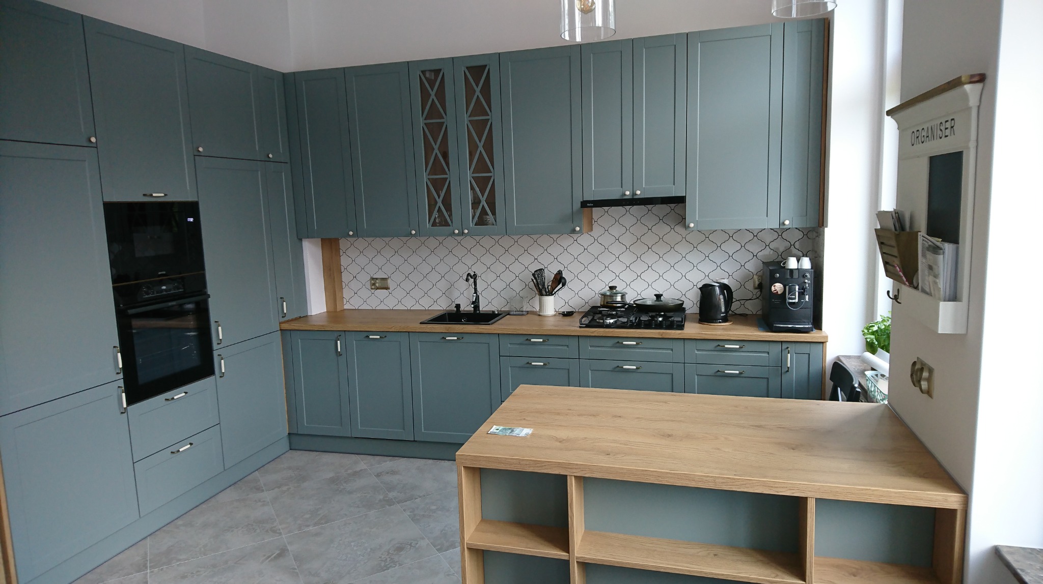 Kompletna kuchnia na wymiar w odcieniach szarości z drewnianym blatem i wyspą, widoczny piekarnik w zabudowie, płyta gazowa, ekspres do kawy i dekoracyjna mozaika nad blatem.