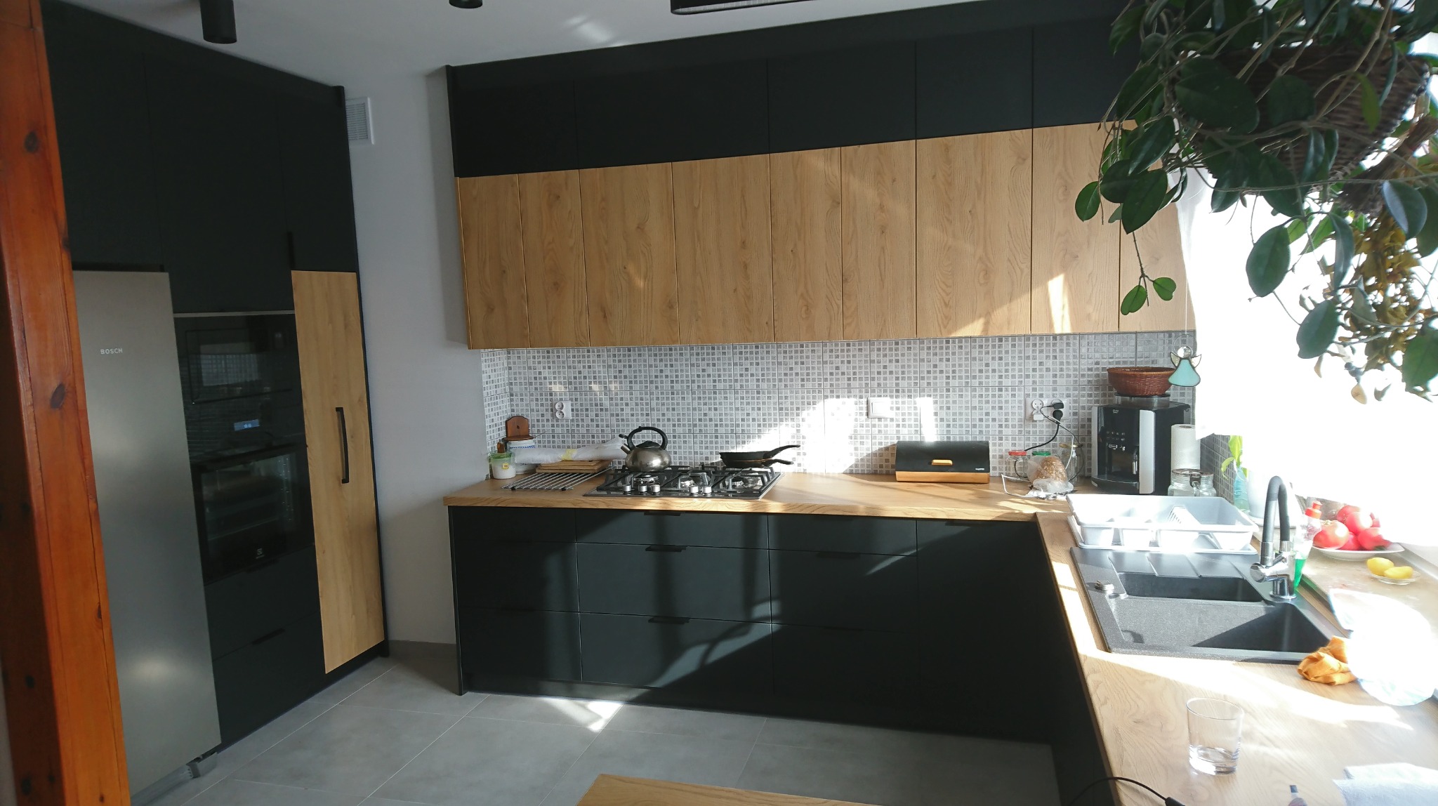 Nowoczesna kuchnia z ciemnymi szafkami dolnymi i drewnianymi górnymi, wykończona mozaiką na ścianie, z widocznym sprzętem AGD marki Bosch i wiszącą rośliną doniczkową.