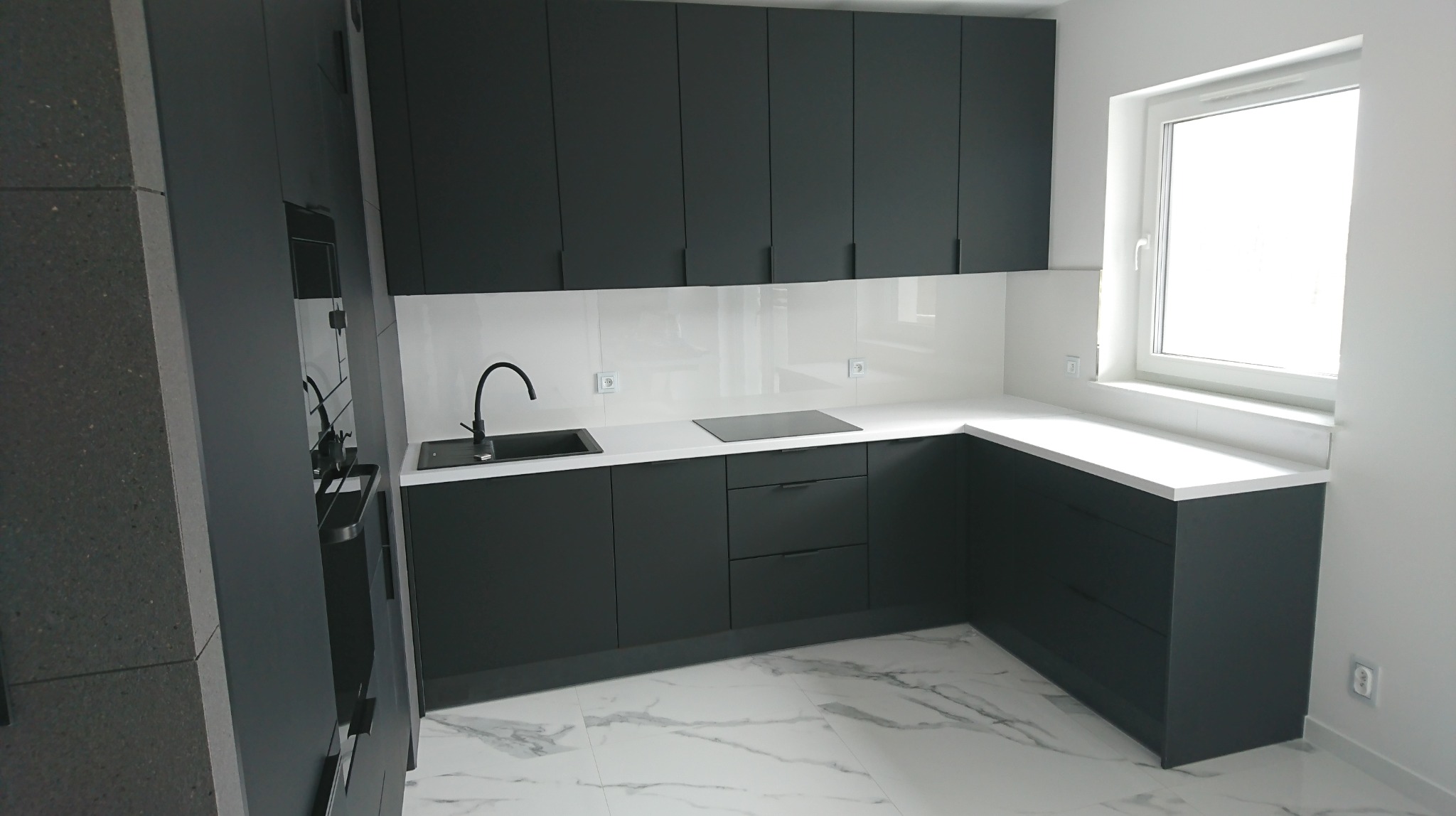 Nowoczesna kuchnia z ciemnymi, matowymi frontami szafek, białym blatem i backsplashiem, marmurowa podłoga, widok na czarny zlew i płytę indukcyjną.
