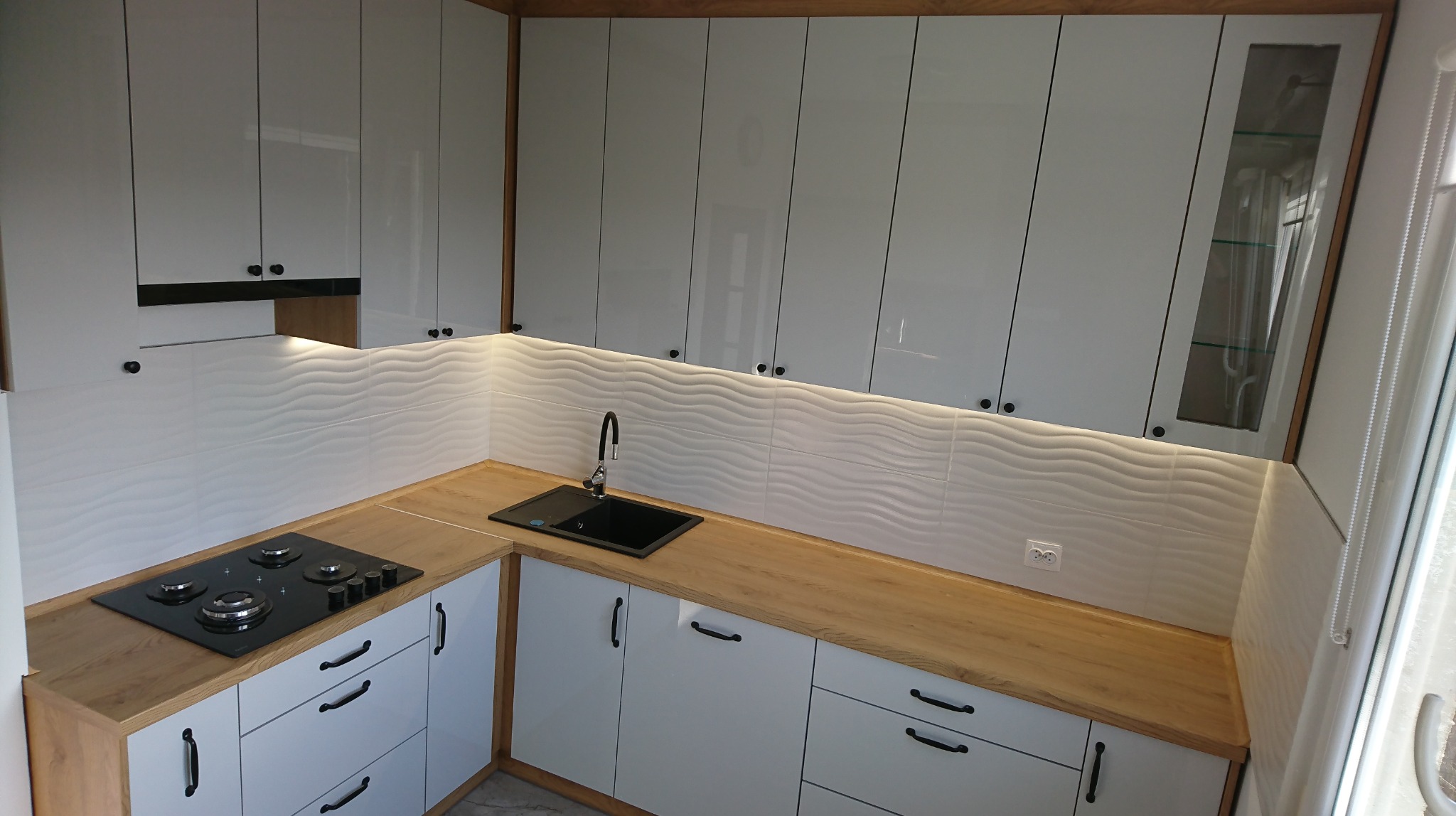 Nowoczesna kuchnia z białymi, lakierowanymi frontami szafek, drewnianym blatem i falistymi płytkami ściennymi podświetlonymi LED, z czarną płytą grzewczą i zlewem.