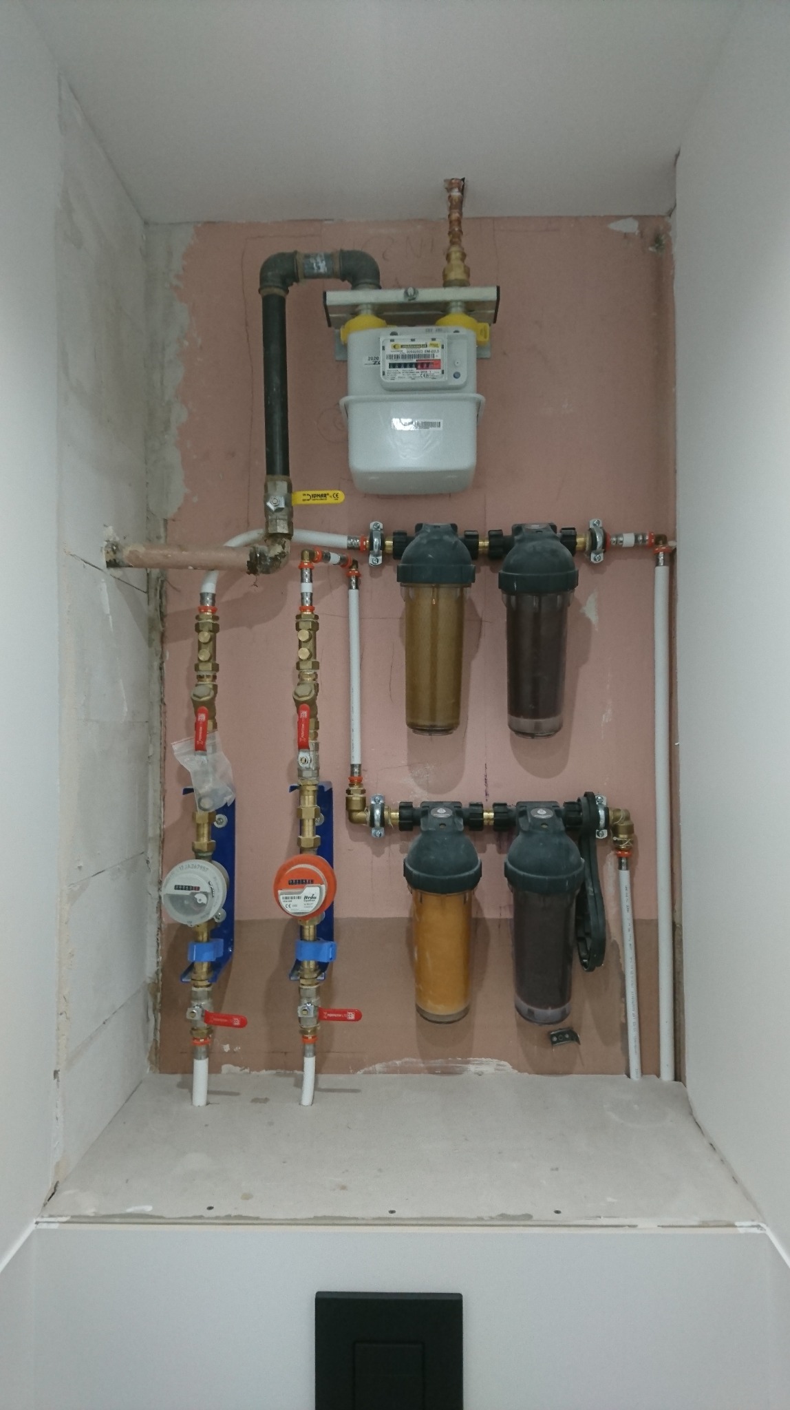 Instalacja wodno-kanalizacyjna i gazowa w trakcie prac wykończeniowych: liczniki, filtry wody i rury w małej wnęce, widoczne niedoskonałości ścian.