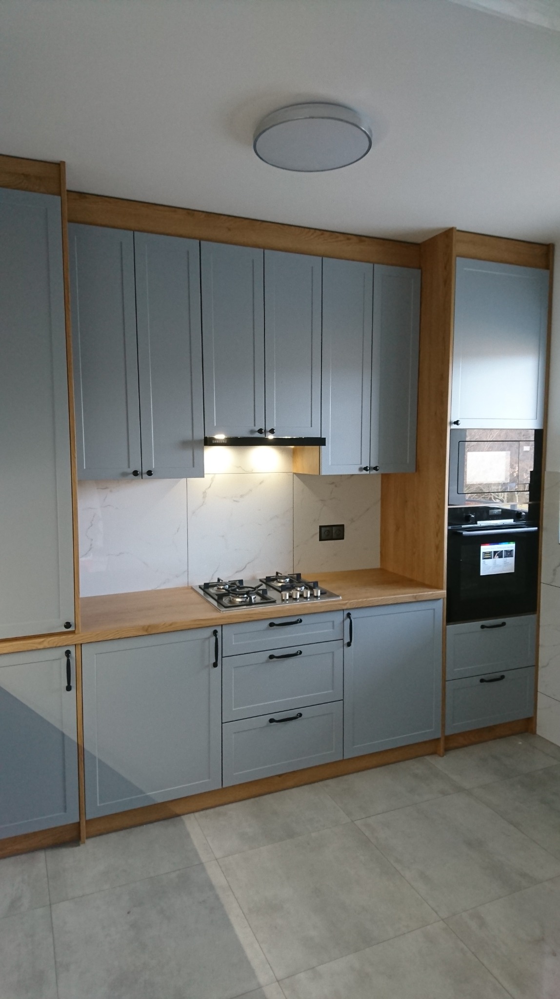 Szare szafki kuchenne z drewnianymi akcentami i blatem, płyta gazowa, marmurowe płytki na ścianie, oświetlenie podszafkowe i czarny piekarnik z mikrofalówką.