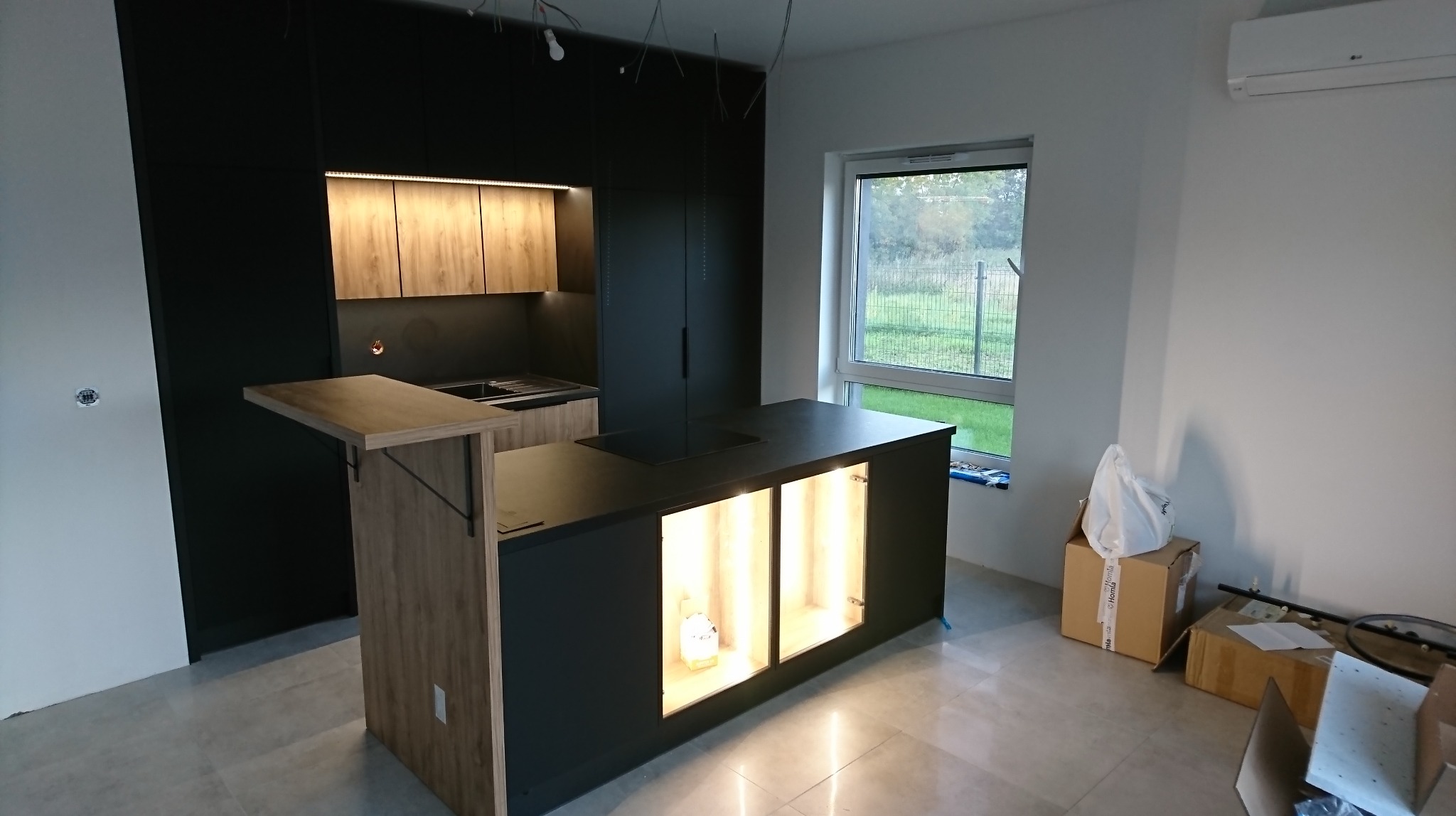 Nowoczesna kuchnia z czarnymi frontami i drewnianym blatem, wyspa z podświetlanymi półkami, widoczne niedokończone prace wykończeniowe.
