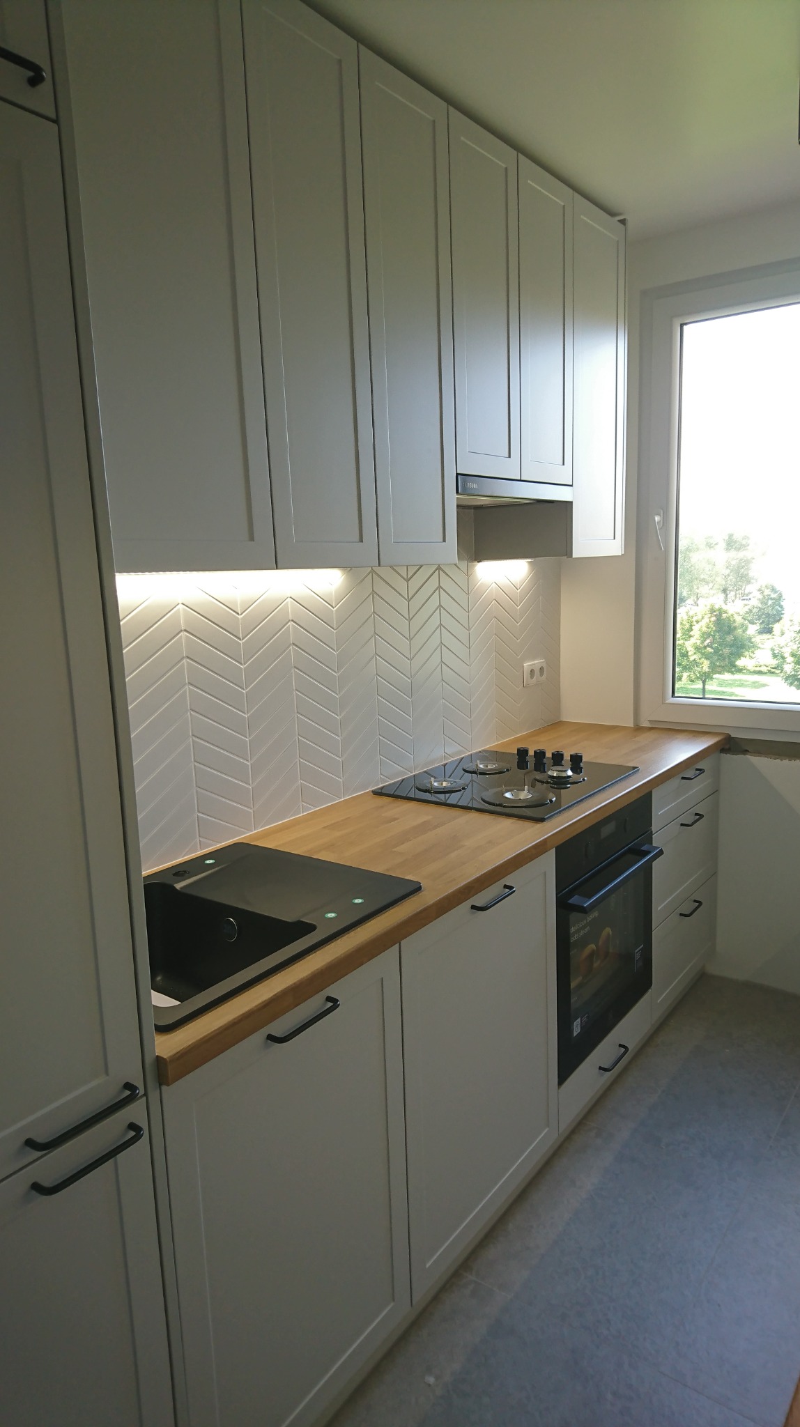 Jasna kuchnia z drewnianym blatem, czarnym zlewem i płytą indukcyjną; białe płytki ułożone w jodełkę na ścianie między szafkami, oświetlenie podszafkowe.