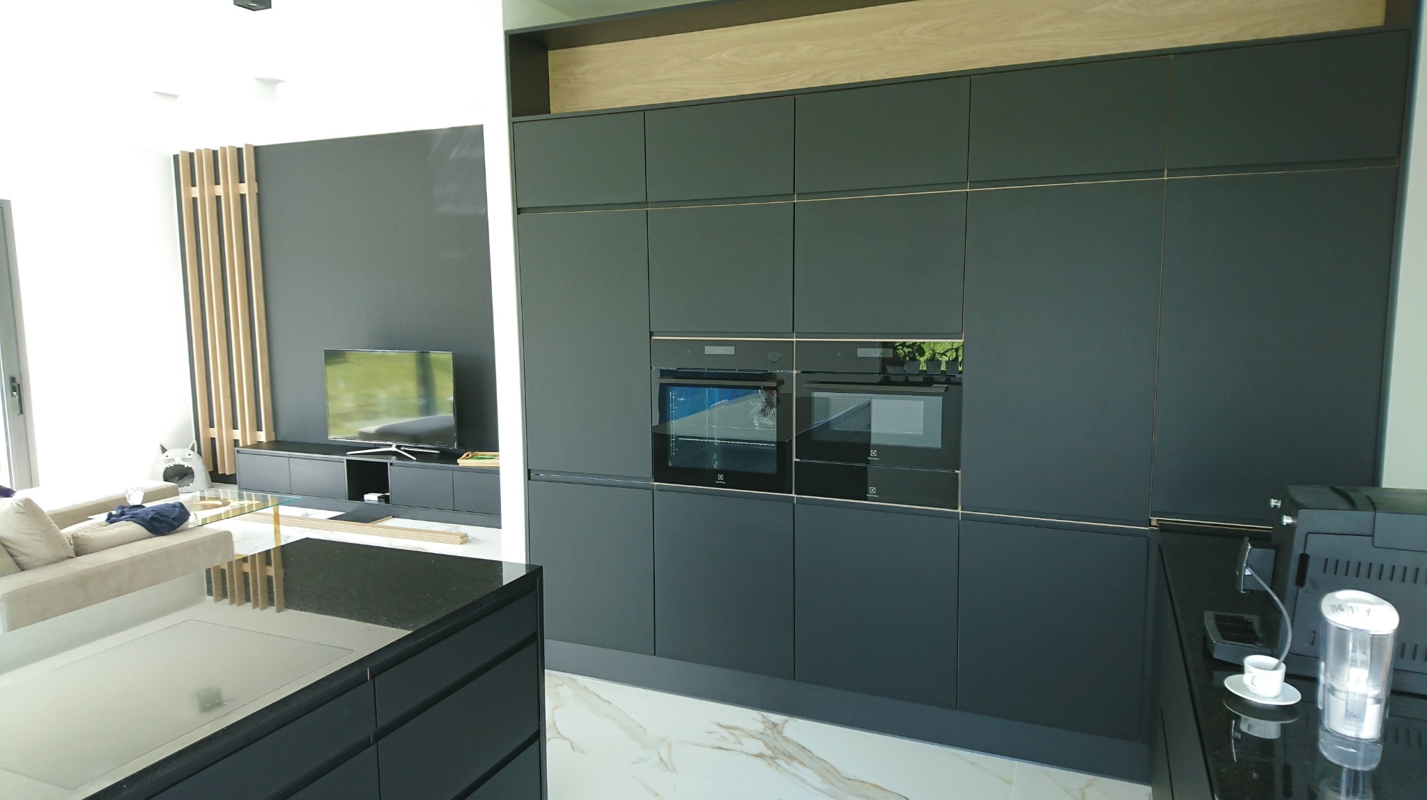 Nowoczesna kuchnia z zabudową w kolorze grafitowym, zintegrowane piekarniki Electrolux, marmurowa podłoga, widok na salon z drewnianymi lamelami i telewizorem.