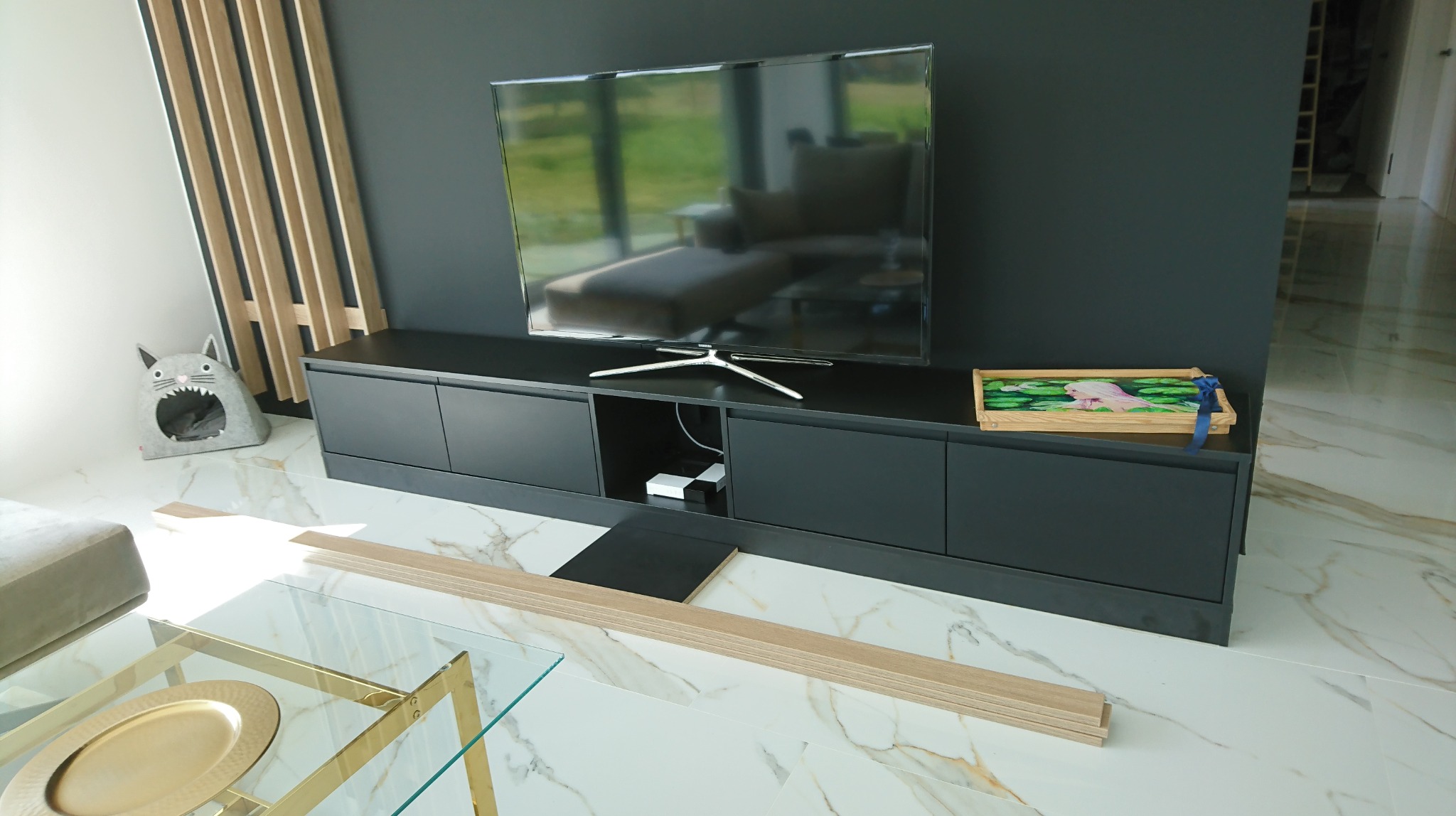 Nowoczesna szafka RTV w kolorze grafitowym z blatem, na której stoi telewizor i ozdobna szkatułka z motywem lilii wodnych, w tle ściana z drewnianymi lamelami, na marmurowej podłodze leżą panele...