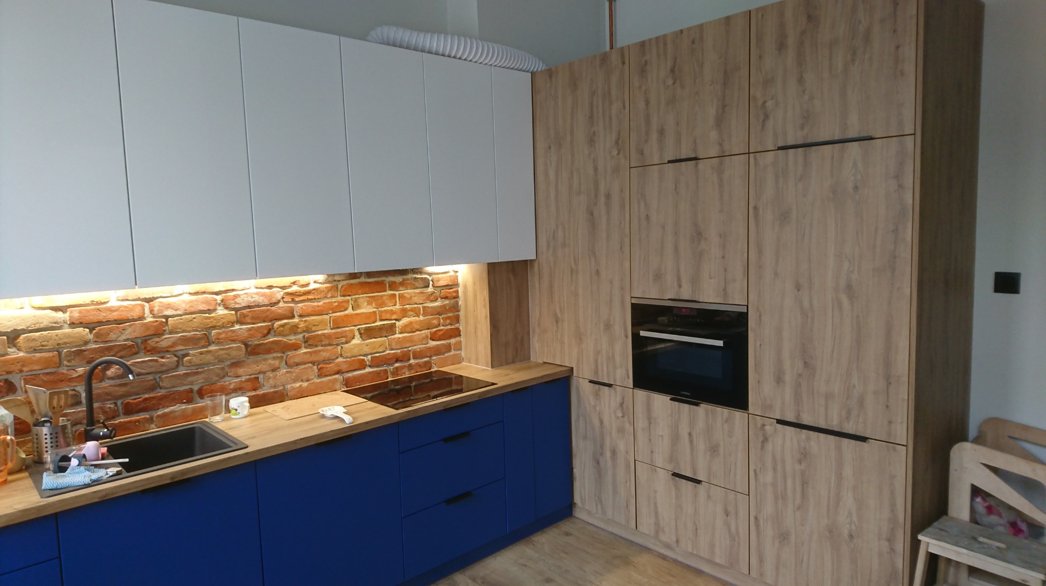 Nowoczesna kuchnia z białymi szafkami górnymi, niebieskimi szafkami dolnymi i ścianą z czerwonej cegły. Szafa w zabudowie z piekarnikiem w kolorze drewna.