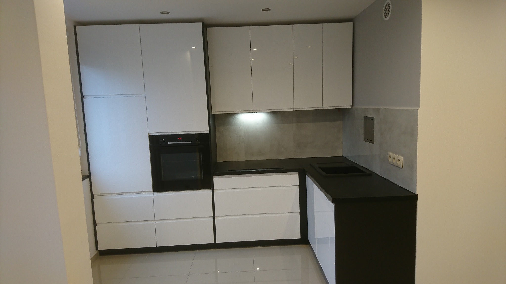 Nowoczesna kuchnia na wymiar z białymi frontami i czarnym blatem, widoczny piekarnik w zabudowie i szare płytki na ścianie.