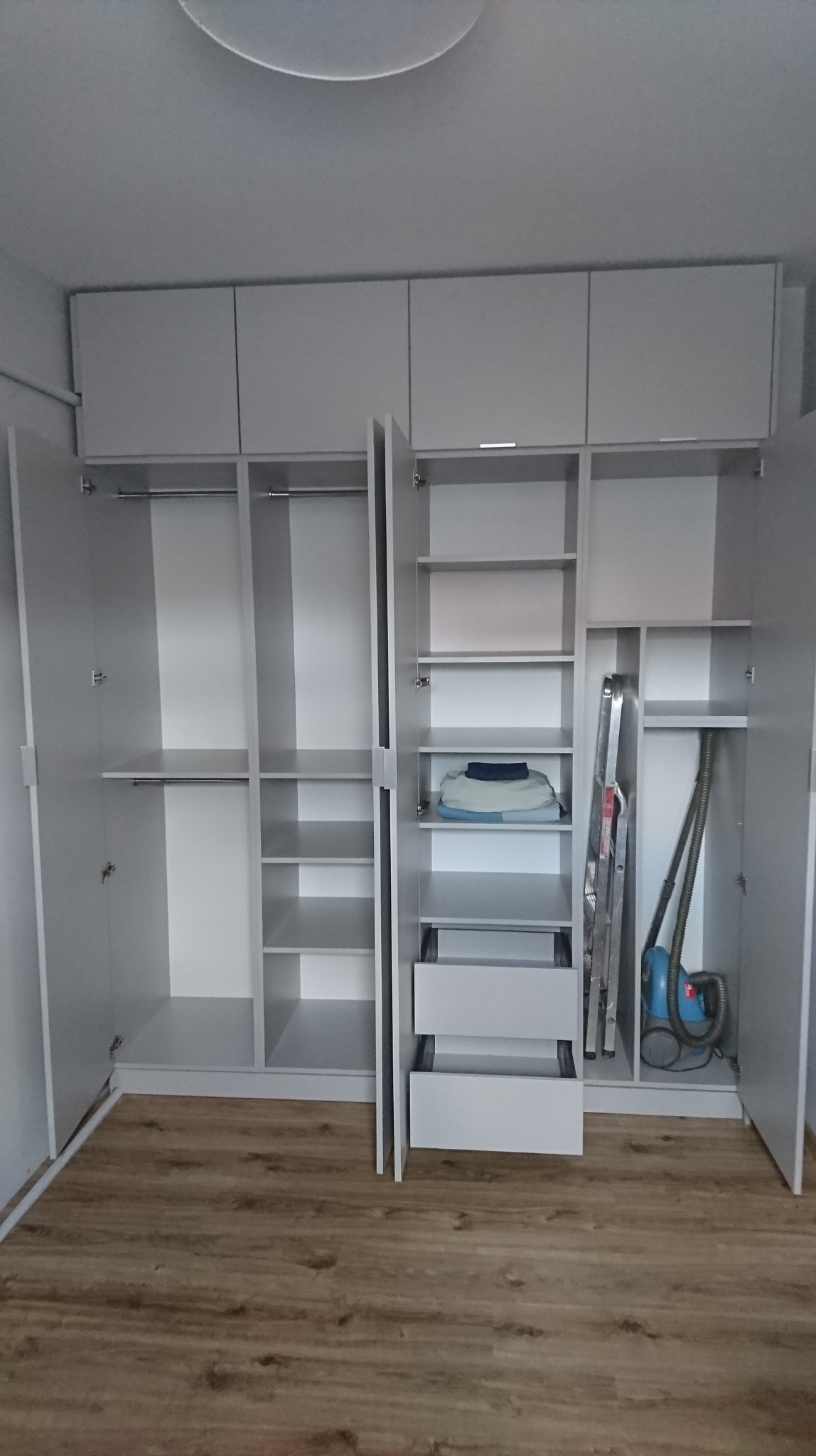 Jasnoszara szafa z otwartymi drzwiami, widoczne półki, szuflady, drążki na ubrania, drabina i odkurzacz wewnątrz.