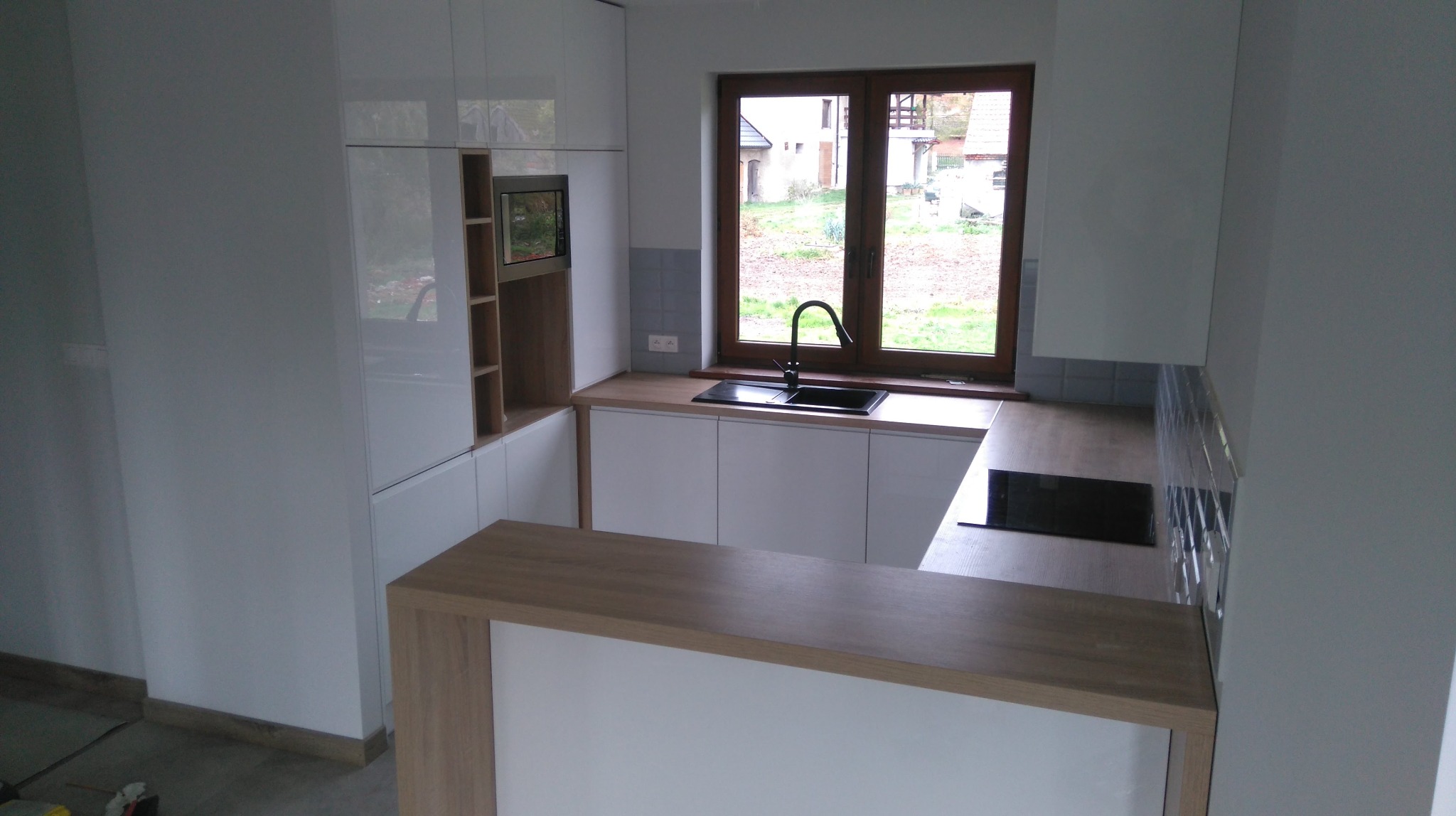 Nowoczesna kuchnia z białymi, lakierowanymi frontami, drewnianym blatem i barkiem, czarnym zlewozmywakiem pod oknem z widokiem na ogród.