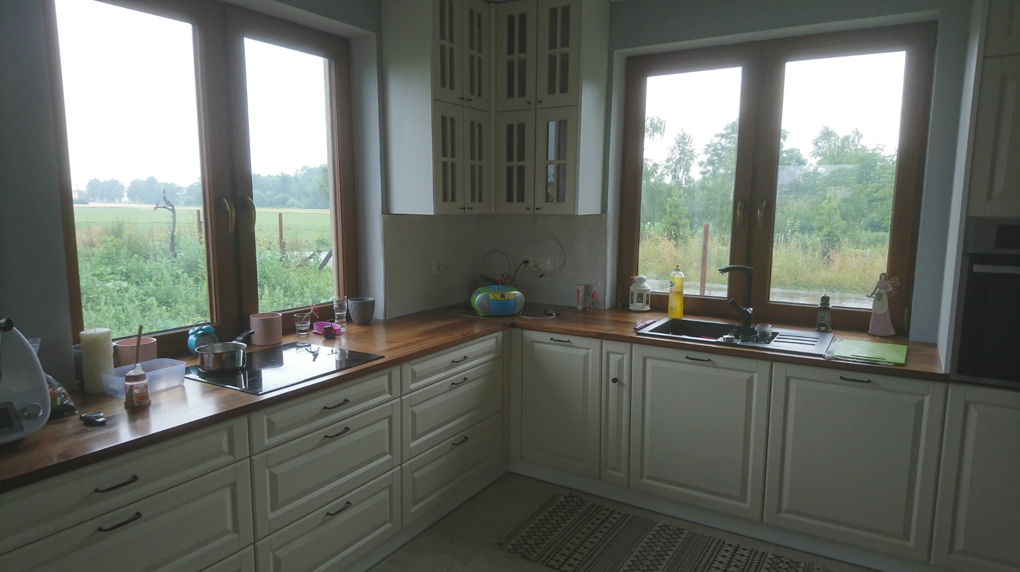 Jasna kuchnia z drewnianym blatem, widok na zielony krajobraz za oknem, na blacie kuchennym stoją różne akcesoria i dekoracje.