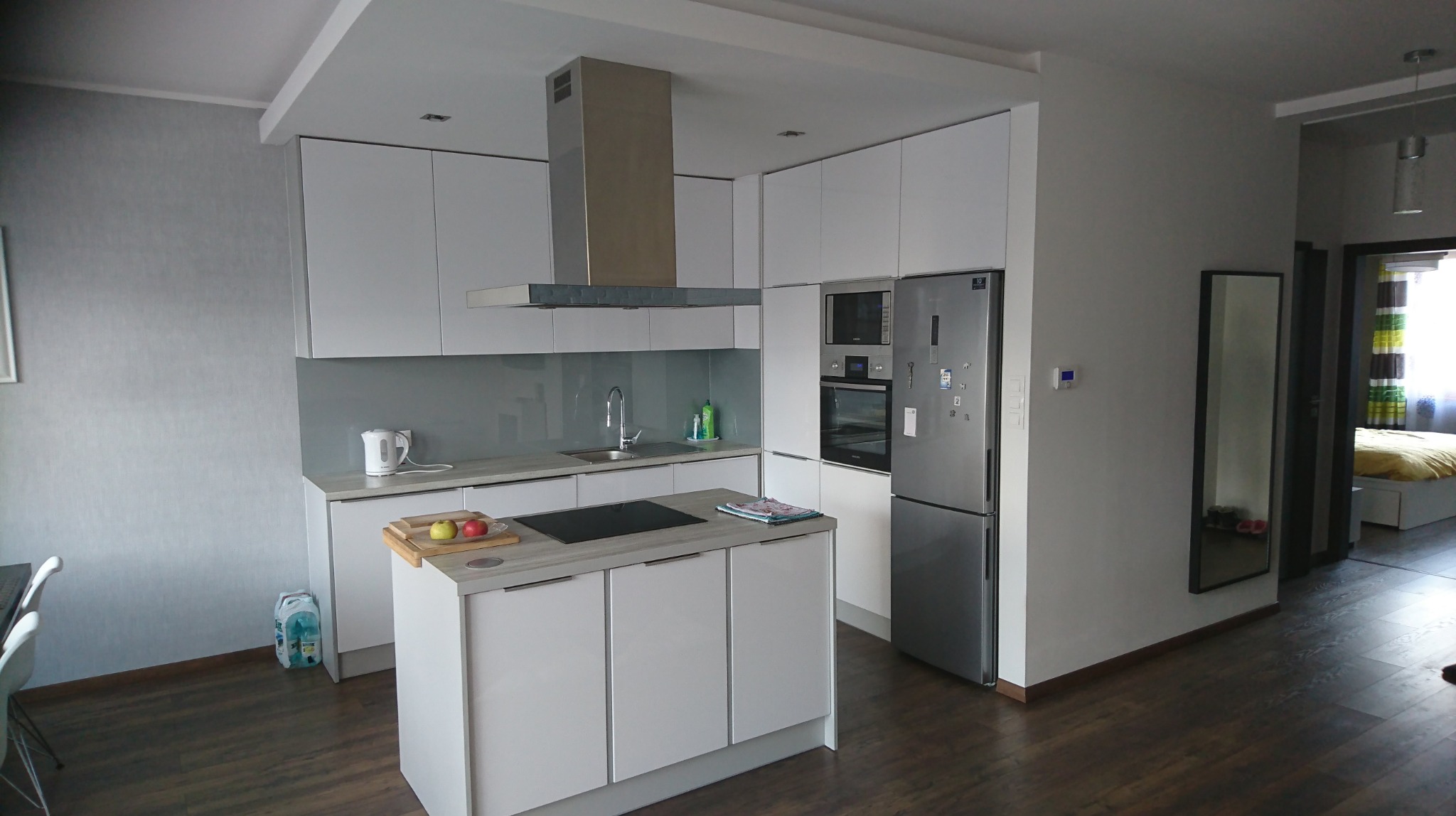 Nowoczesna, minimalistyczna kuchnia z białymi frontami, wyspą i szarym blatem, widziana z perspektywy wejścia do pokoju, z widoczną lodówką i okapem.