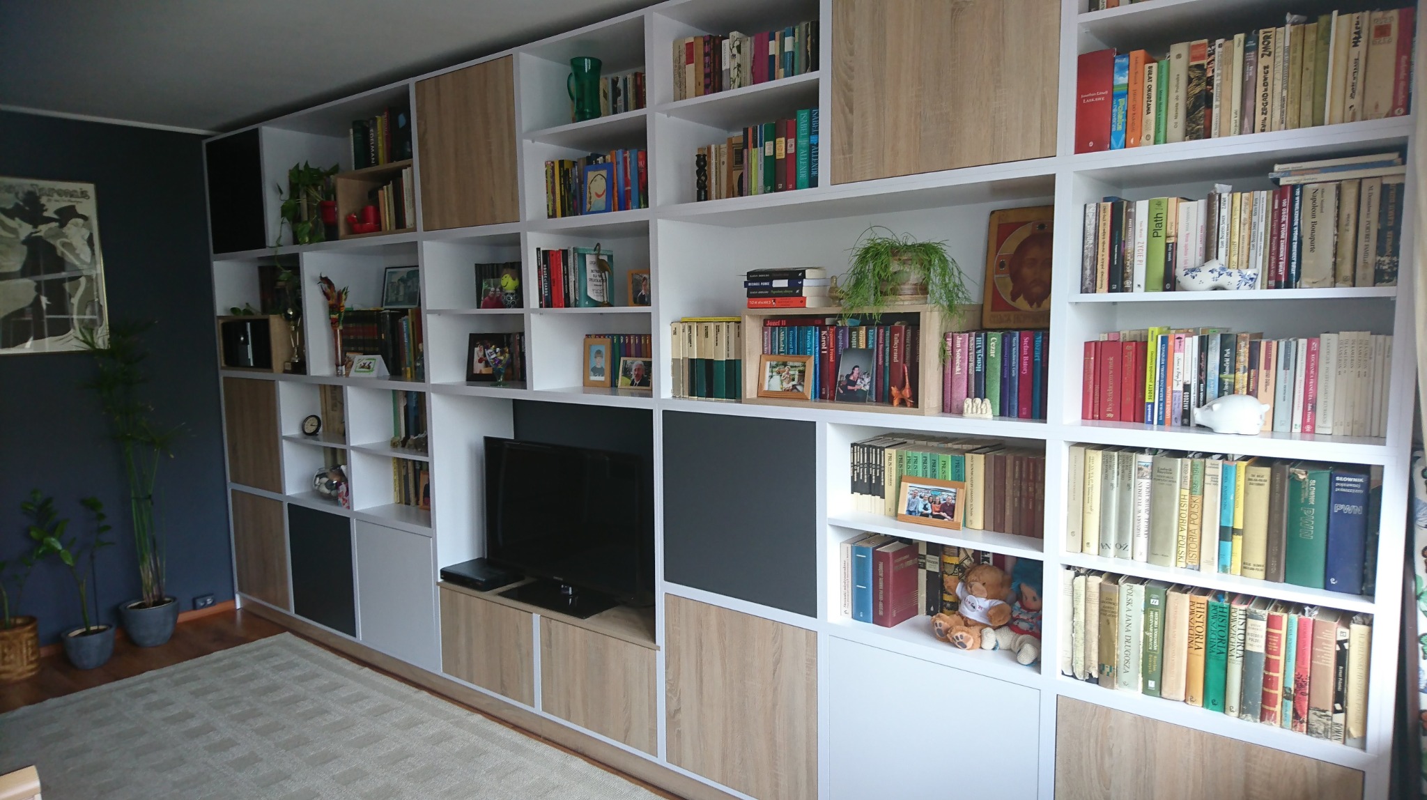 Duża, biała szafa na wymiar z półkami, wypełniona książkami, dekoracjami i telewizorem w salonie z ciemną ścianą w tle.