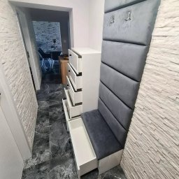 STOLARZ TOMASH TOMASZ WIŚNIEWSKI - Przedpokój z białą komodą z wysuniętymi szufladami, szarą tapicerowaną ścianką z siedziskiem i wieszakami, wykończony płytkami imitującymi kamień i czarną marmurową podłogą.