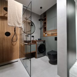 STOLARZ TOMASH TOMASZ WIŚNIEWSKI - Nowoczesna łazienka z drewnianymi akcentami: kabina prysznicowa typu walk-in z drewnianą ścianą, okrągłe lustro, umywalka nablatowa, pralka pod blatem, wisząca toaleta w kolorze czarnym, szare...
