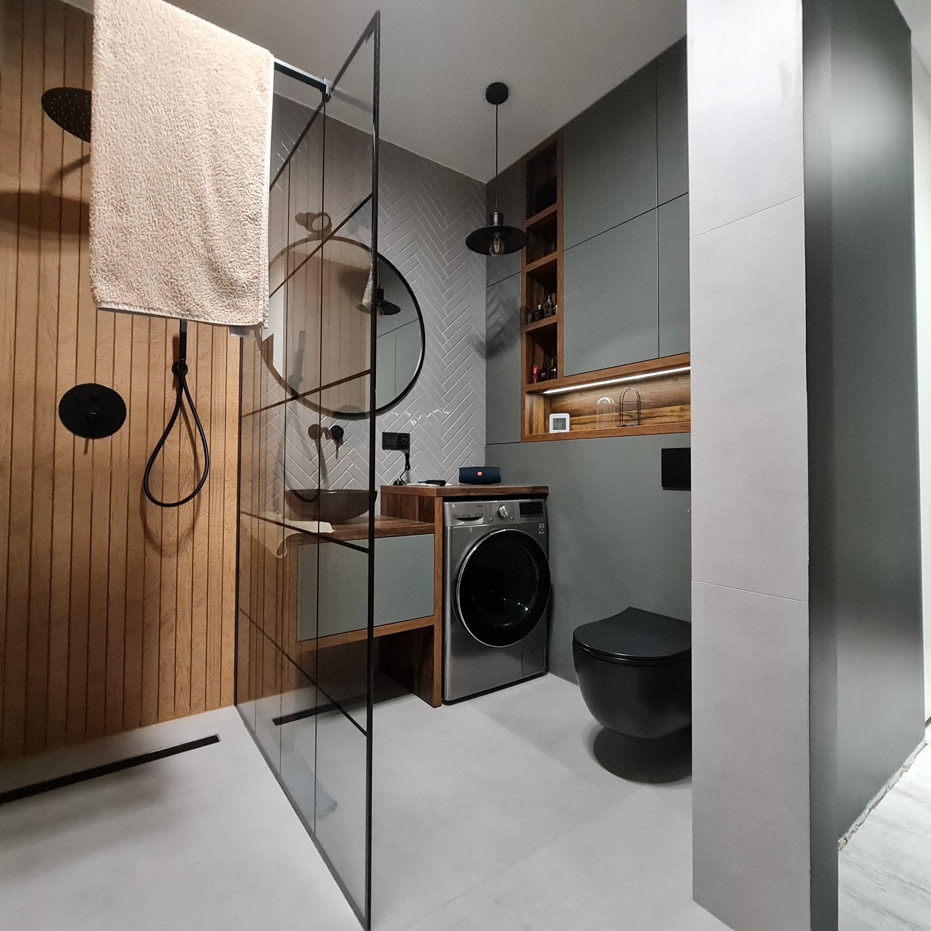 Nowoczesna łazienka z drewnianymi akcentami: kabina prysznicowa typu walk-in z drewnianą ścianą, okrągłe lustro, umywalka nablatowa, pralka pod blatem, wisząca toaleta w kolorze czarnym, szare...
