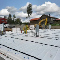 Przygotowany fundament pod dom z instalacją ogrzewania podłogowego, widoczna żółta koparka Komatsu i zabudowania w tle, dzień słoneczny.