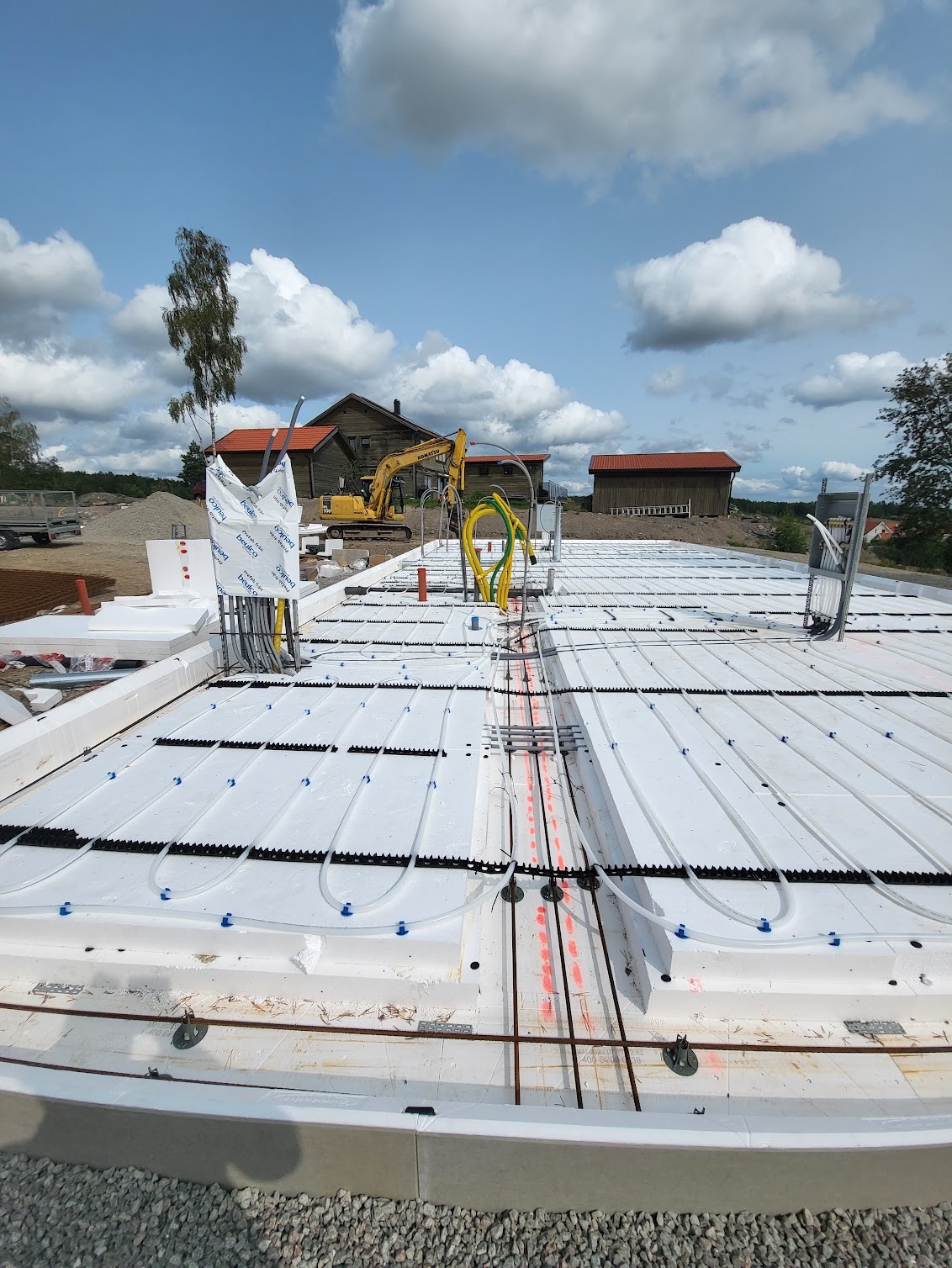 Przygotowanie płyty fundamentowej z instalacją ogrzewania podłogowego na budowie domu, z widocznym szkieletem zbrojeniowym i izolacją termiczną, w tle budynek i koparka.
