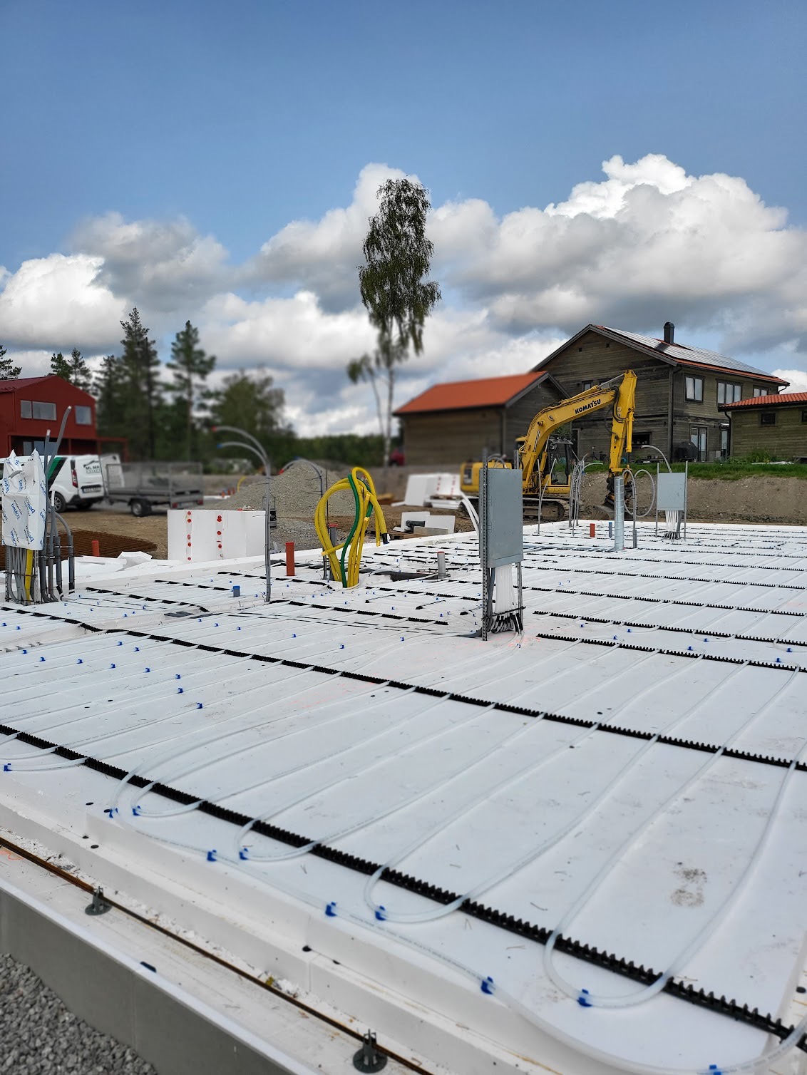 Przygotowany fundament pod dom z instalacją ogrzewania podłogowego, widoczna żółta koparka Komatsu i zabudowania w tle, dzień słoneczny.
