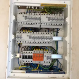 Rozdzielnia elektryczna w białej obudowie, widoczne liczne przewody, bezpieczniki i elementy montażowe. Instalacja po modernizacji, gotowa do użytku.