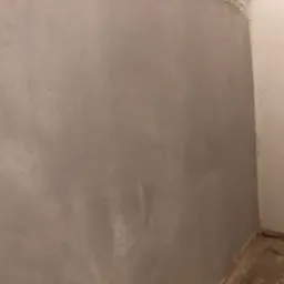 Świeżo położona, szara gładź na ścianie w rogu pomieszczenia, z widocznymi śladami po szpachli. Druga ściana pomalowana na biało, widoczne niedoskonałości w dolnej części.