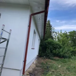 Biała elewacja domu z brązowymi rynnami i podbitką, widok z dołu, drabina oparta o ścianę. Świeżo otynkowana ściana i prace wykończeniowe wokół budynku.