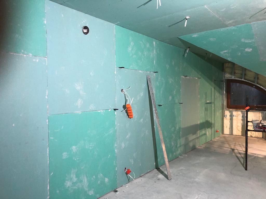 Montaż zielonych płyt gipsowo-kartonowych na ścianach i suficie poddasza ze skośnym oknem. Widoczne profile i instalacja elektryczna. W tle rusztowanie.