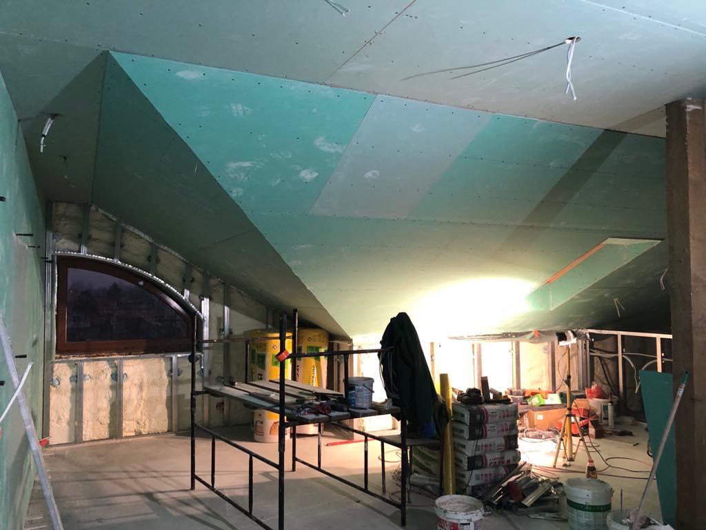 Poddasze z oknem w trakcie zabudowy płytami G-K w kolorze zielonym. Widoczne rusztowanie, materiały budowlane i izolacja. Skosy sufitowe i ściany częściowo obłożone.