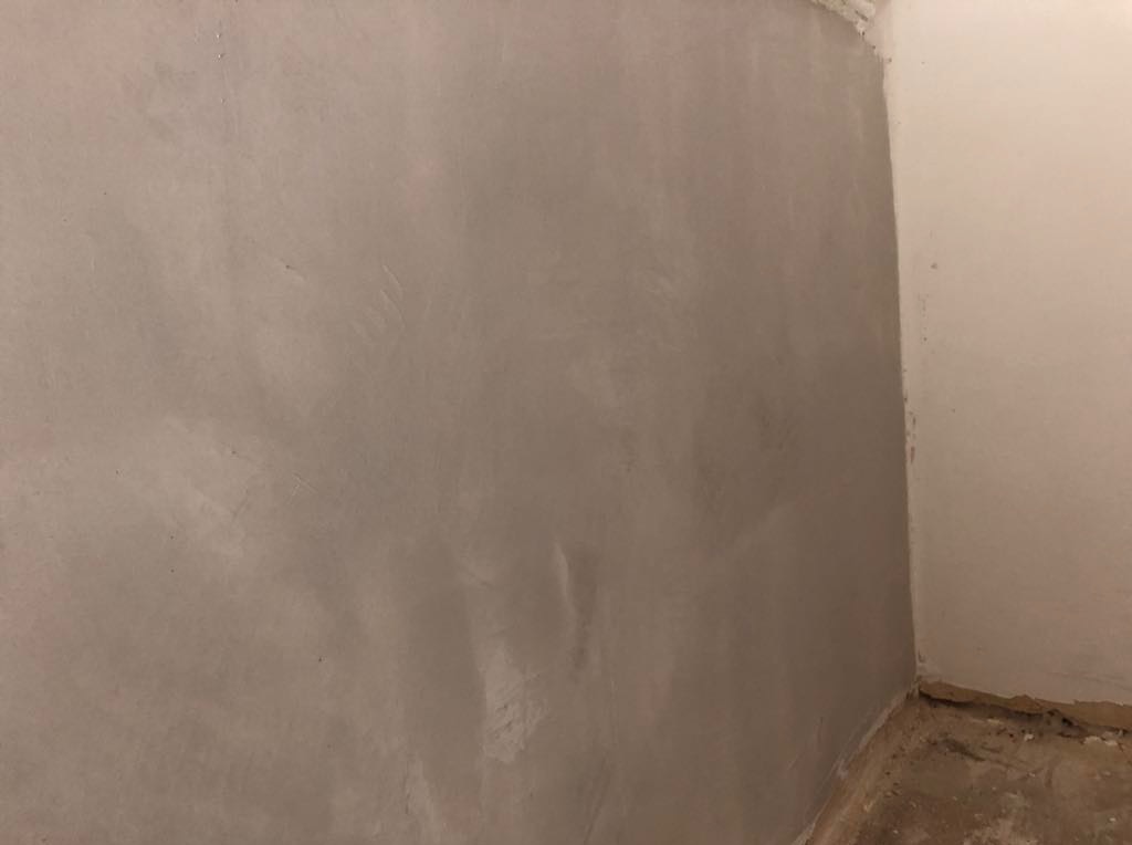 Świeżo położona, szara gładź na ścianie w rogu pomieszczenia, z widocznymi śladami po szpachli. Druga ściana pomalowana na biało, widoczne niedoskonałości w dolnej części.