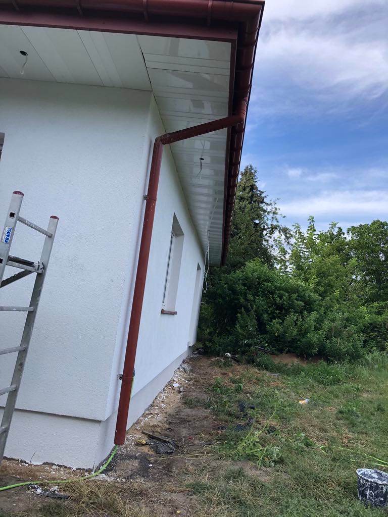 Biała elewacja domu z brązowymi rynnami i podbitką, widok z dołu, drabina oparta o ścianę. Świeżo otynkowana ściana i prace wykończeniowe wokół budynku.